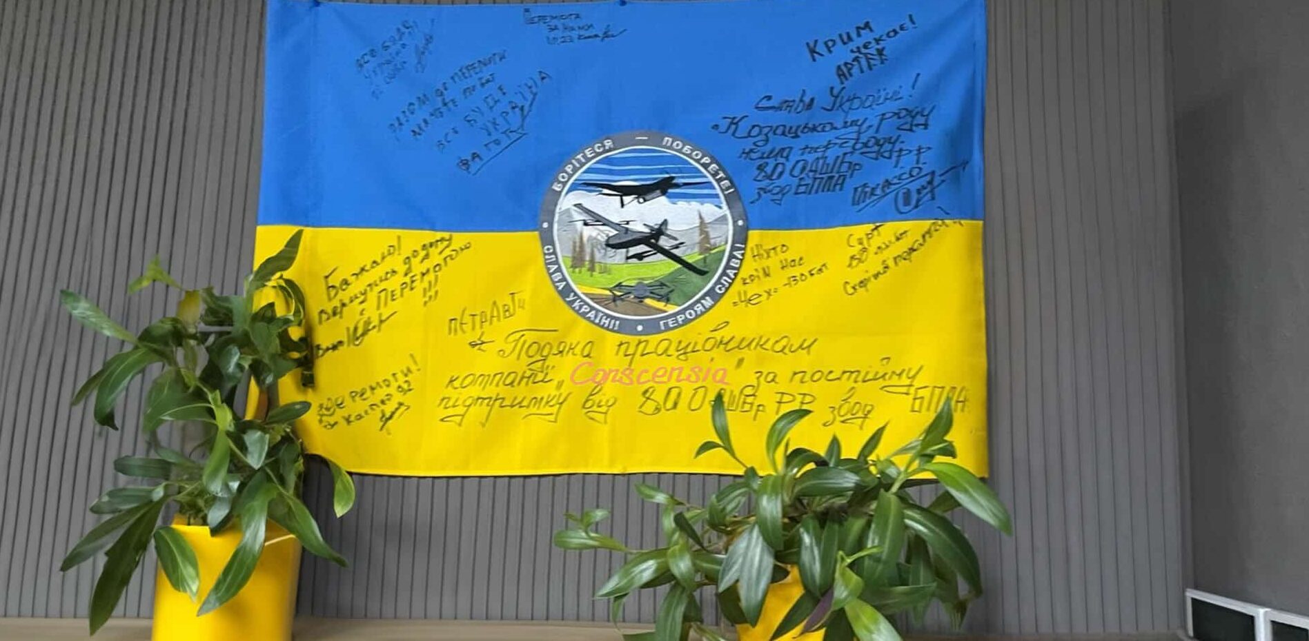 Et blåt og gult ukrainsk flag med signaturer og en besked på ukrainsk, der udtrykker taknemmelighed til Conscensias ansatte for deres støtte, hængende på en væg.